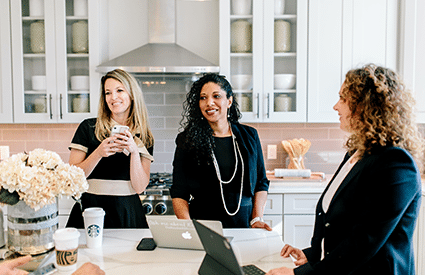 three female real estate agents dressed professionally in black in white kitchen with laptops open and coffee cups on the counter