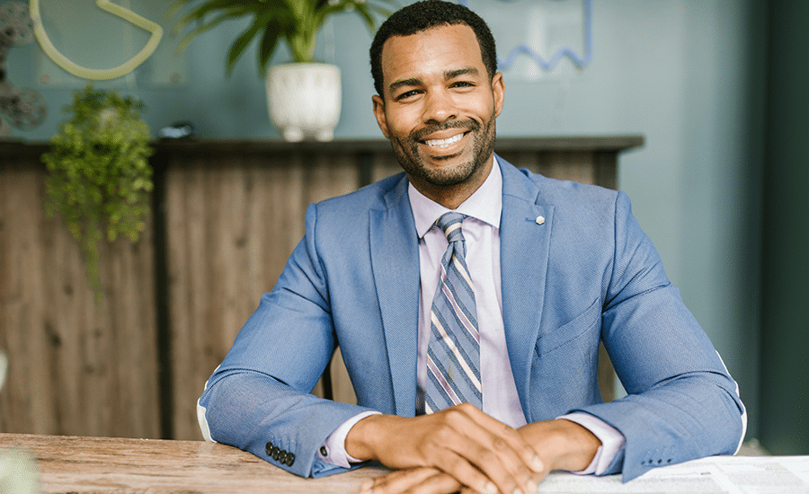 african american man in light blue suit smiling at office table with green plants