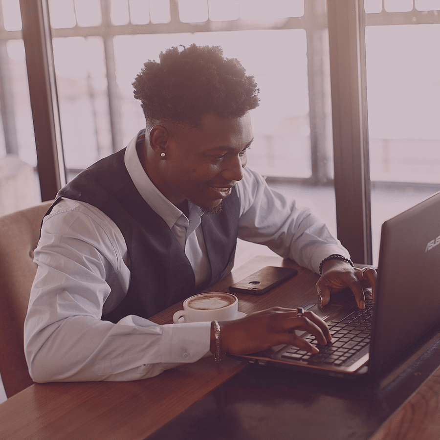 african american man in vest and button down shirt smiling while typing on computer with coffee and cell phone on desk