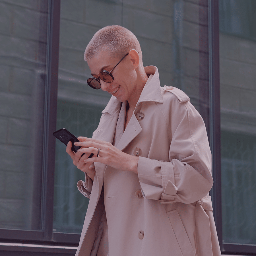 caucasian female with buzzed blonde hair cut with big sunglasses and coat smiling at phone while walking in city