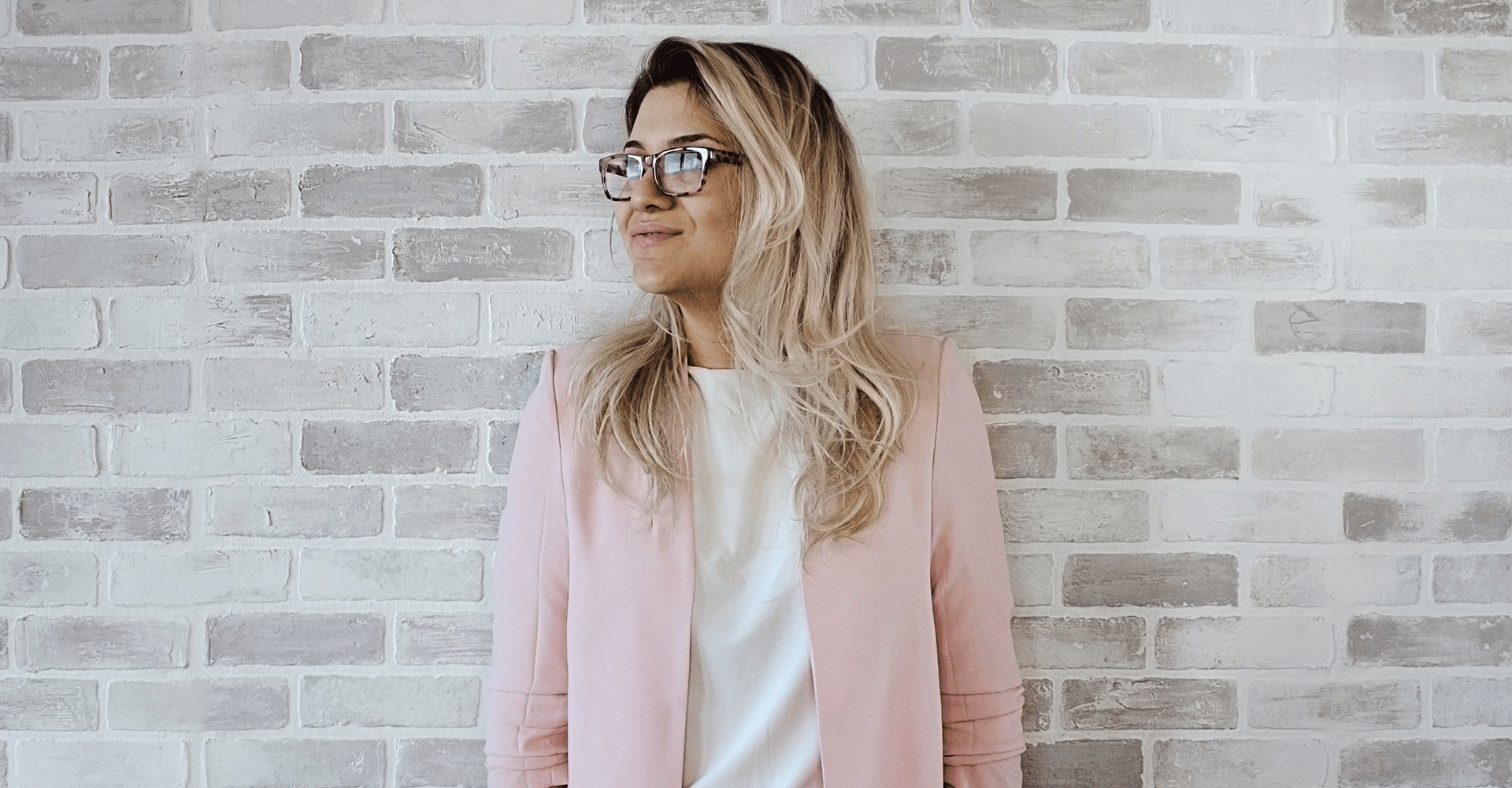 millennial woman with blonde hair wearing pink jacket leaning against light brick wall