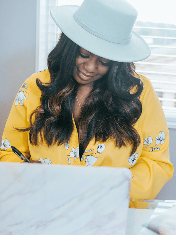 millennial woman with dark hair wearing bright yellow blouse and white hat writing notes