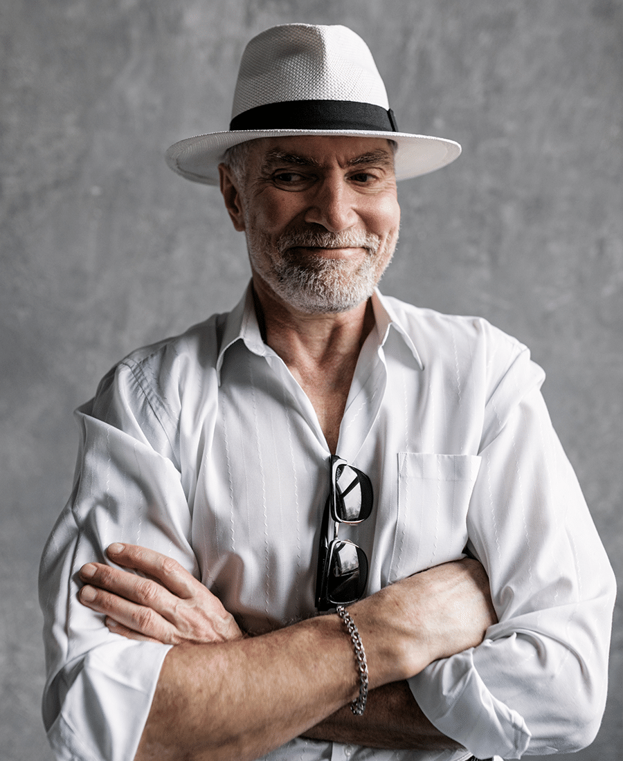 older caucasian male with gray beard grinning wearing white fedora with arms crossed