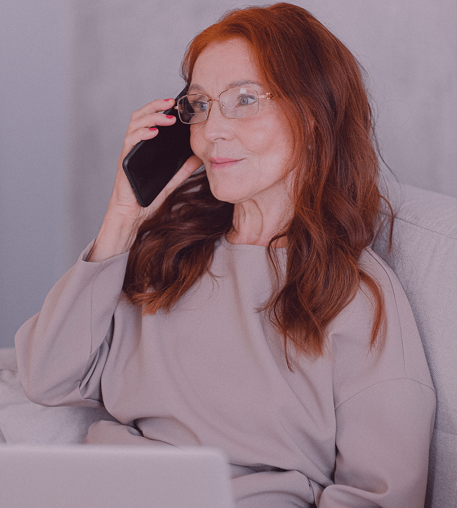 older woman with red hair and glasses sitting on couch calling on phone