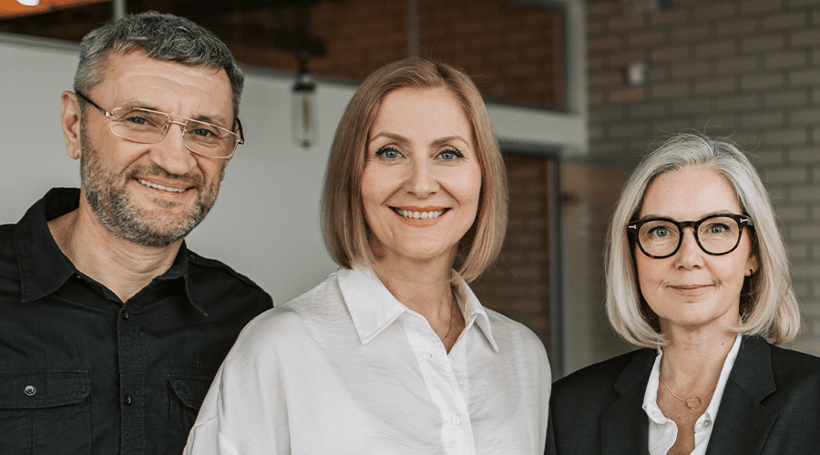 one man with gray hair and beard with glasses and two women with short hair smiling in office