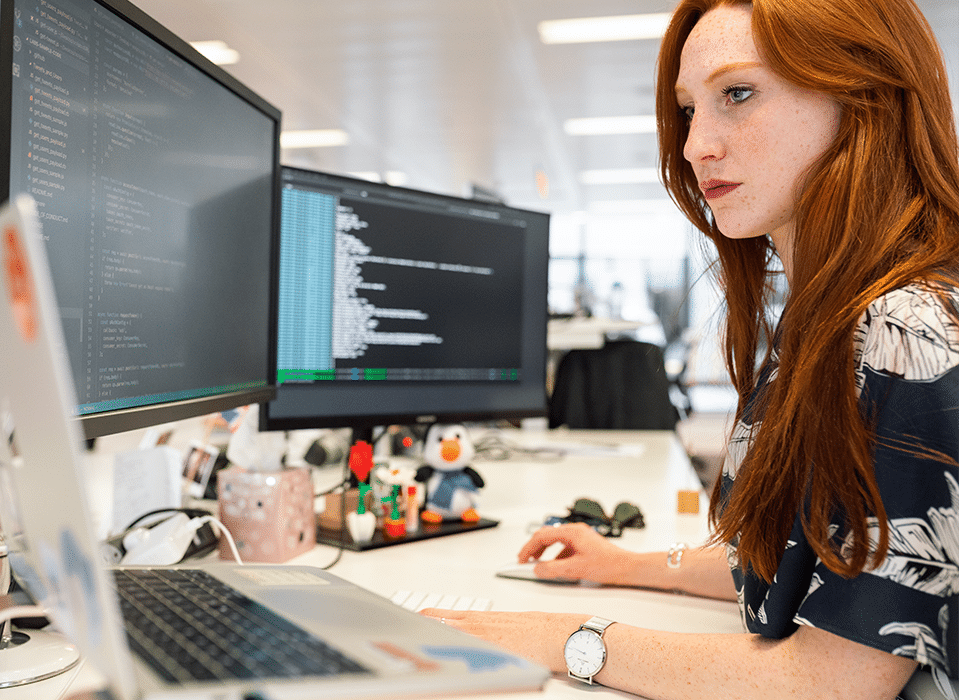 red haired woman in office looking at three computer screens