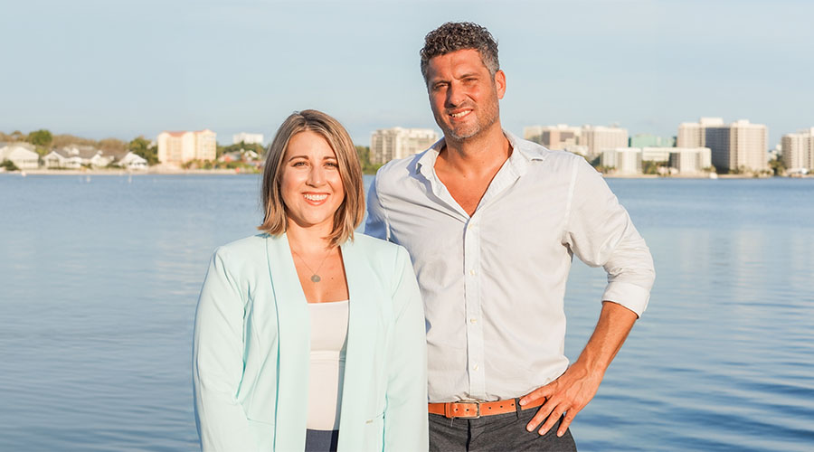 white woman and man real estate team dressed in professional coastal pastel colors along bay with homes and condos along horizon